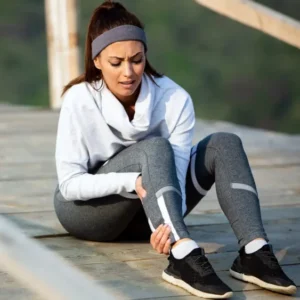 Vrouw in sportkleding zit op een houten brug en wrijft over haar onderbeen, mogelijk door stijve spieren na inspanning.