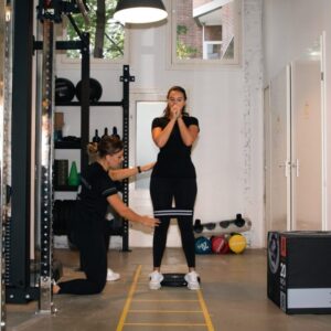Fysiotherapeut begeleidt jonge vrouw na een voorste kruisband blessure operatie - zij doet een spring en balansoefening in een oefenzaal. van F3 Fysio, met sportmateriaal en loopladder op de vloer.