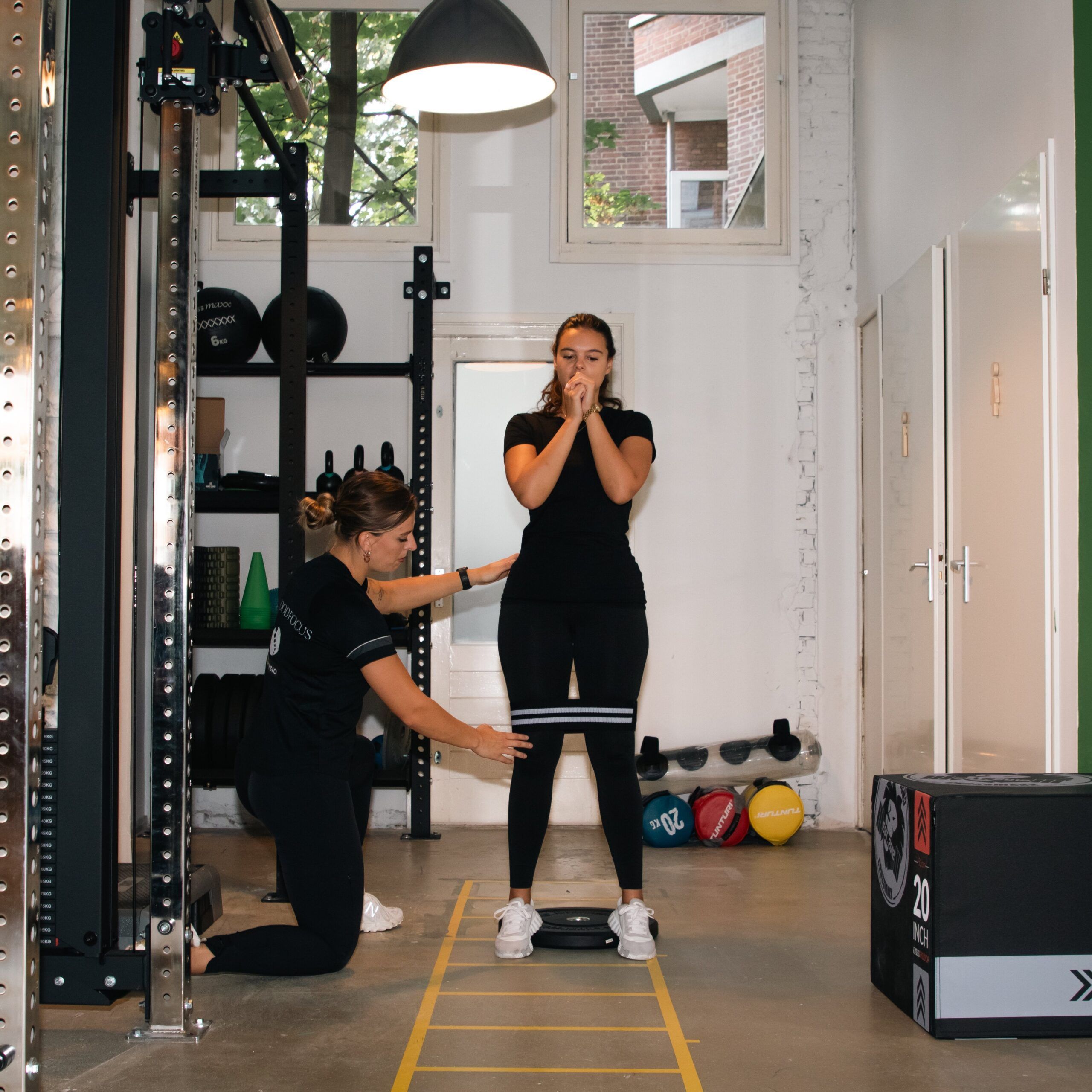 F3 Fysio Rotterdam - Noord, Fysiotherapeut begeleidt jonge vrouw tijdens balans- en krachttraining in de oefenzaal van F3 Fysio, met sportmateriaal en loopladder op de vloer.