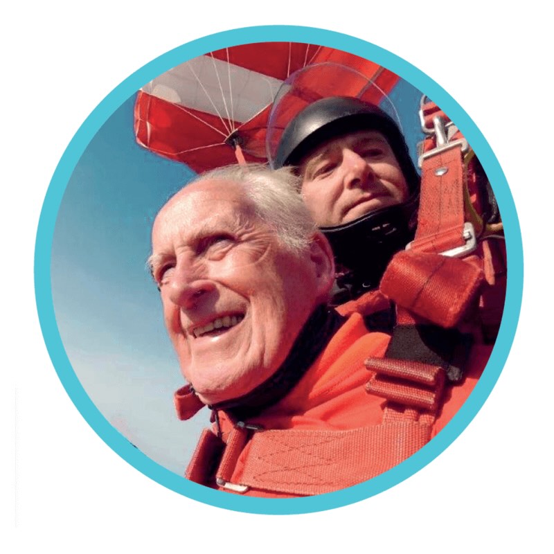 Boele, een 85-jarige man, geniet van een tandemsprong tijdens een parachutesprong. Met een brede glimlach en in een rood skydivepak zweeft hij door de lucht, veilig vastgegespt aan zijn instructeur, met de parachute al open op de achtergrond. Zijn gezicht straalt plezier en avontuur uit.