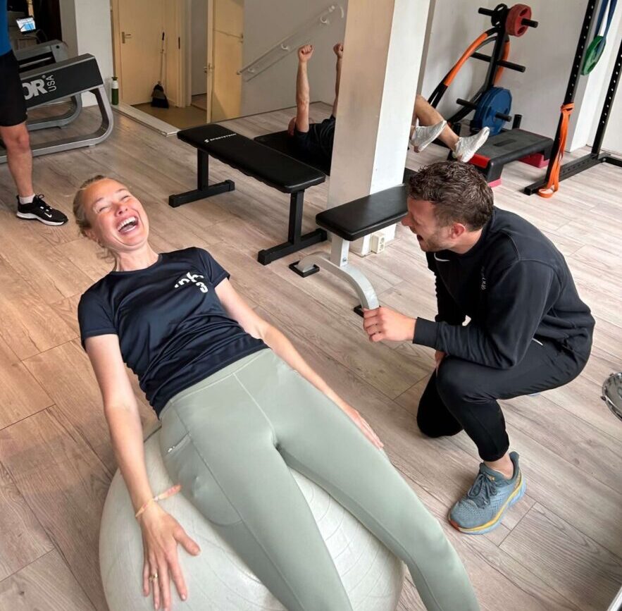 Een vrouw ligt lachend op een fitnessbal terwijl een fysiotherapeut haar begeleidt. De praktijkruimte heeft fitnessapparatuur en gewichten op de achtergrond
