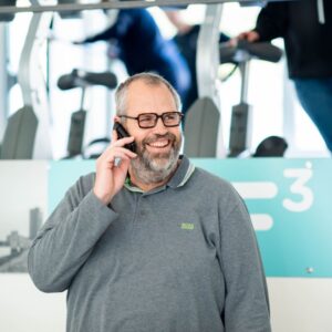 Een lachende man in de trainingsruimte van F3 Fysio, pratend aan de telefoon met fitnessapparatuur op de achtergrond. Creëert een vriendelijke en actieve sfeer die past bij een professionele fysiotherapie- en trainingsomgeving.