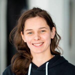 Een lachende jonge vrouw draagt een zwarte hoodie met logo in een professionele omgeving. De afbeelding straalt energie en positiviteit uit, ideaal voor thema's over gezondheid, fitness en welzijn.