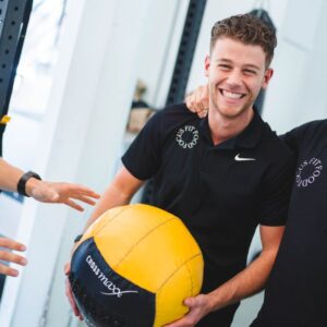 Lachende man met een gele trainingsbal in een crossfit-sportschool, met gewichtentrainingsapparatuur op de achtergrond, ter promotie van gezondheid en conditie.