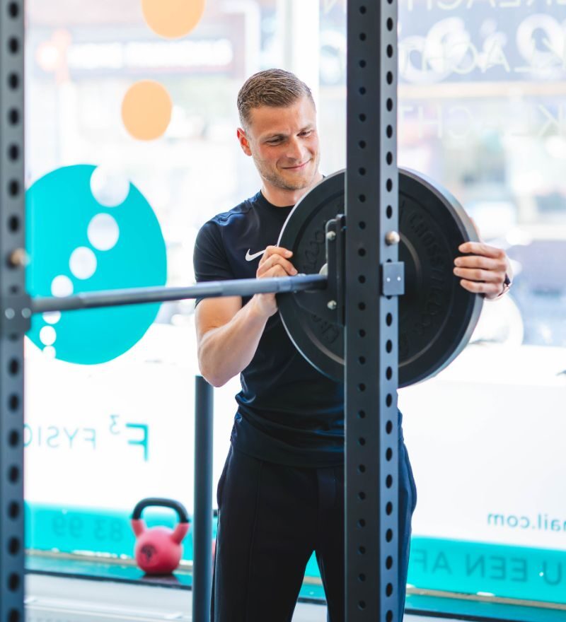 Lachende man die een gewichtsschijf aan het verstellen is in een moderne fysio met kleurrijk versierde ramen op de achtergrond. Perfect voor wie op zoek is naar fitness of fysieke training.
