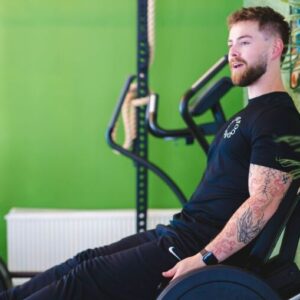 Een man traint zijn knieën met gewichten, zelfs voor een goed verlichte groene muur. Hij draagt ​​zwarte sportkleding en heeft een verzorgd haar en een baard.