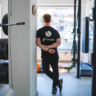 F3 fysiotherapeut staat in lichte fitnessruimte en kijkt naar client die oefening doet op een yogamat